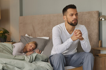 Young man deep in thoughts while his wife sleeps in the background