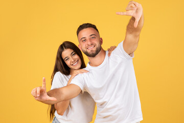 Glad young european man and woman in white t-shirts making frame with hands, capturing a moment