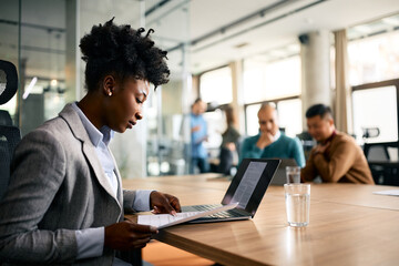 Black businesswoman analyzing reports while using laptop in office.