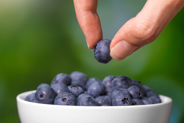 Close-up of female fingers pulling out a ripe blueberry berry from a white bowl on a blurry green background, macro photography