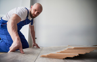Man in work overalls installing laminate floor in apartment under renovation. Male construction worker laying laminate timber flooring in living room with white wall on background.