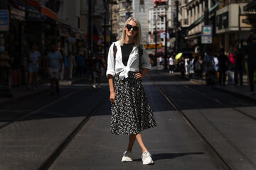 Obraz premium Beautiful blonde young woman in sunglasses poses on one of the main streets of Istanbul on a sunny day. Stylish female tourist with a backpack in Turkey