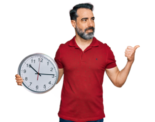 Middle aged man with beard holding big clock pointing thumb up to the side smiling happy with open mouth