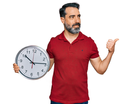Middle aged man with beard holding big clock pointing thumb up to the side smiling happy with open mouth
