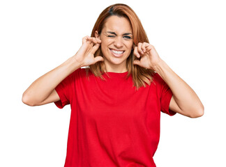 Hispanic young woman wearing casual red t shirt covering ears with fingers with annoyed expression for the noise of loud music. deaf concept.