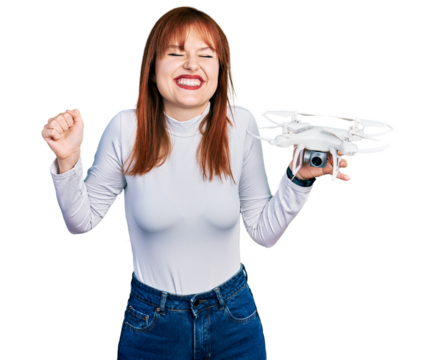 Redhead young woman holding drone screaming proud, celebrating victory and success very excited with raised arm