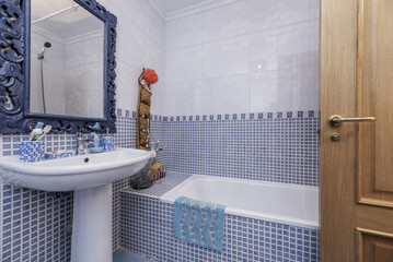 A bathroom with blue and white tiles, a dividing border, a white bathtub, a mirror with a blue...