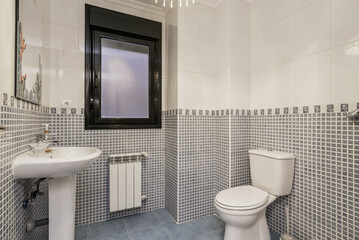 A bathroom with stoneware and white tiles, a dividing border, porcelain sink, matching toilet and black aluminum and glass window