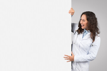 Cheerful young female doctor peeking from behind blank white placard board for text or design, free...