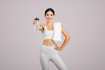 Smiling slim caucasian lady in sportswear with towel showing bottle of water, isolated on grey...