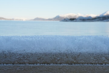 Schneeschicht auf Holzgeländer vor Meereskulisse und Bergen mit Schnee in Norwegen, Eiskristalle, Raureif, 