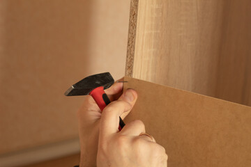A man hammers a nail into a piece of wood. Assembling furniture at home. The photo shows hands and a hammer.