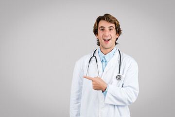 Fototapeta premium Joyful man doctor pointing to side, stethoscope around neck on grey backdrop
