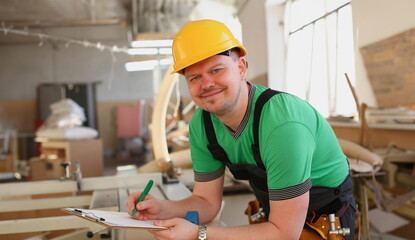 Portrait of young attractive man in work clothes and yellow helmet crossed arms smiling at shop for manufacturing furniture and details home interior