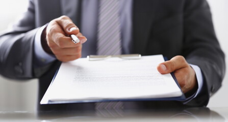 Male arm in suit offer contract form on clipboard pad and silver pen to sign closeup. Strike a...