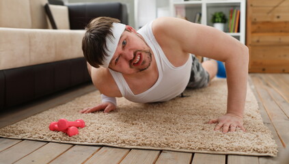 A young handsome man with excess weight makes the first fitness training at home on the carpet is wrung out and he ery heavily loaded does not stimulate to lose weight. - obrazy, fototapety, plakaty