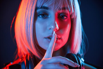 Gesture of be quiet, finger is by the lips. Woman with white hair is in studio with neon colors