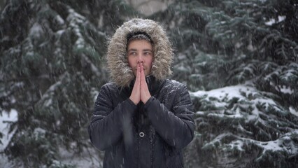 A young man stands against a background of fir trees under heavy snowfall and rubs his hands...
