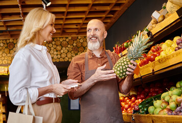 jolly mature salesman with beard showing pineapple to his joyful female customer by grocery stall - Powered by Adobe