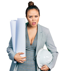Young hispanic girl holding safety helmet and blueprints in shock face, looking skeptical and...