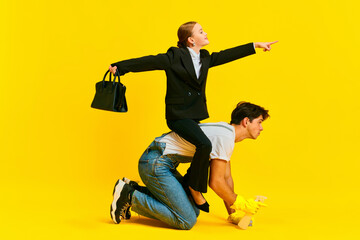 School girl dressed like boss sits on back of adult man, janitor, and shows him where to go against vivid yellow background.