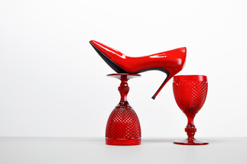 Red wine glasses and a red women's high heel shoe on a white table and background. Balancing...