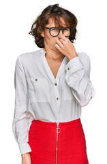 Young hispanic woman wearing business style and glasses smelling something stinky and disgusting,...
