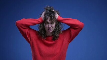 Frustrated young woman pulling her hair in anxiety and desperation on blue backdrop with red sweater. Anxious 20s female person