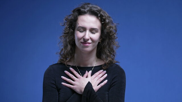 Grateful woman puts hands on chest feeling heartwarmed, smiling expression of hopeful and faithful 20s person, feeling thankful