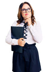 Young beautiful caucasian woman wearing business shirt and glasses holding folder serious face thinking about question with hand on chin, thoughtful about confusing idea