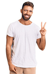 Young hispanic man wearing casual white tshirt smiling with happy face winking at the camera doing victory sign. number two.