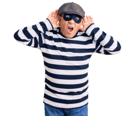 Senior handsome man wearing burglar mask and t-shirt trying to hear both hands on ear gesture, curious for gossip. hearing problem, deaf