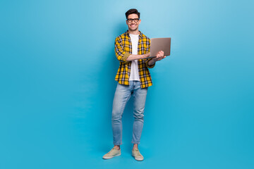 Full size body photo of young guy wear yellow shirt with jeans and going to work using macbook laptop isolated on blue color background - obrazy, fototapety, plakaty