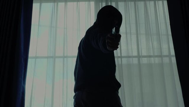 Mysterious man wearing black hoodie holding a pistol wearing a cloth mask covering his face completely in front of the door, Silhouette and dark concept image,threat of firearms,attack or defense.