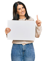 Middle age brunette woman holding blank empty banner surprised with an idea or question pointing finger with happy face, number one