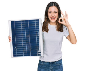 Young beautiful girl holding photovoltaic solar panel doing ok sign with fingers, smiling friendly...