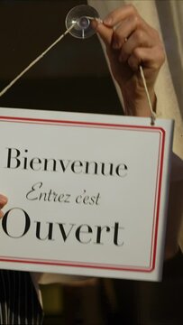 Vertical video of female shop owner turning ouvert ferme sign in French on cafe glass door. Woman staff chef of restaurant turning open closed sign in French.