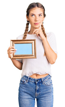 Beautiful caucasian woman with blonde hair holding empty frame serious face thinking about question with hand on chin, thoughtful about confusing idea