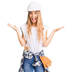 Obraz na płótnie Canvas Beautiful caucasian woman with blonde hair wearing hardhat and painter clothes celebrating victory with happy smile and winner expression with raised hands