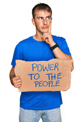 Young blond man holding power to the people banner serious face thinking about question with hand...