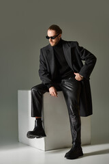 full length, fashionable model in trendy sunglasses and coat sitting on white cube on grey backdrop