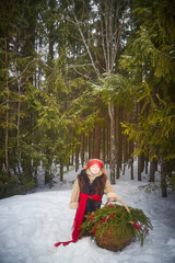 Little girl in thick coat and a red sash with basket of fir branches and red berries in cold winter...