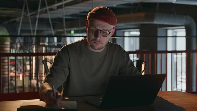 A Young Freelance Man Of 30 Years Old With Glasses Is Sitting Working At A Laptop In A Production Room, In A Coworking. A Male Programmer Writes Important Information From A Computer In A Notebook. 4k