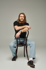 full length of bearded good looking man with long hair sitting in jeans and t-shirt on chair on grey