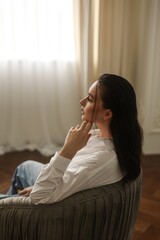 A young woman sits on a chair and looks out the window. A thoughtful girl.