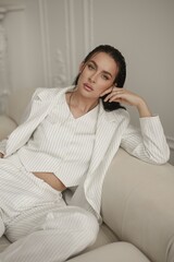 Impressive Caucasian woman in a white pinstripe suit gracefully poses on a small white couch. The clean, minimalist studio setting, with white walls and a parquet flooring