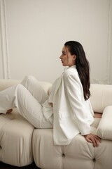 A self-assured and stylish brunette woman in a white pinstripe suit confidently poses on a white couch in a well-lit studio. The backdrop, consisting of white walls and a parquet floor