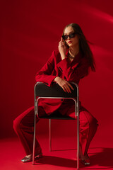 Fashionable confident woman wearing trendy sunglasses, red suit blazer, classic trousers, metallic silver color shoes, sitting on chair, posing on red background. Full-length studio fashion portrait