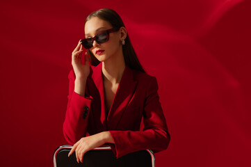 Fashionable confident woman wearing trendy sunglasses, red suit blazer, posing on red background....