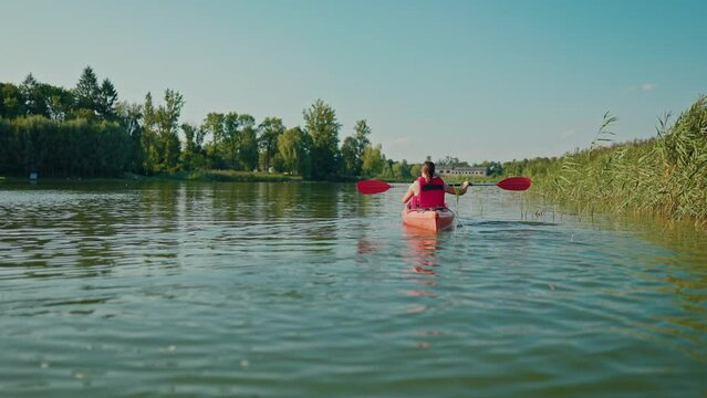View of young woman actively paddling while sitting in red canoe. Rowing with oar near river bank. Having good time during kayaking in landscape with beautiful nature. Relaxation of body.